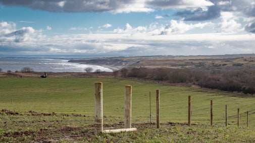 A large green field in the foreground, with a line of new wooden fence posts leading downhill off to the right. The view behind it is of a bay with cliffs and headlands of the Durham Coast beyond, with choppy blue-grey sea on the left. The sky is bright with blue patches between clouds.